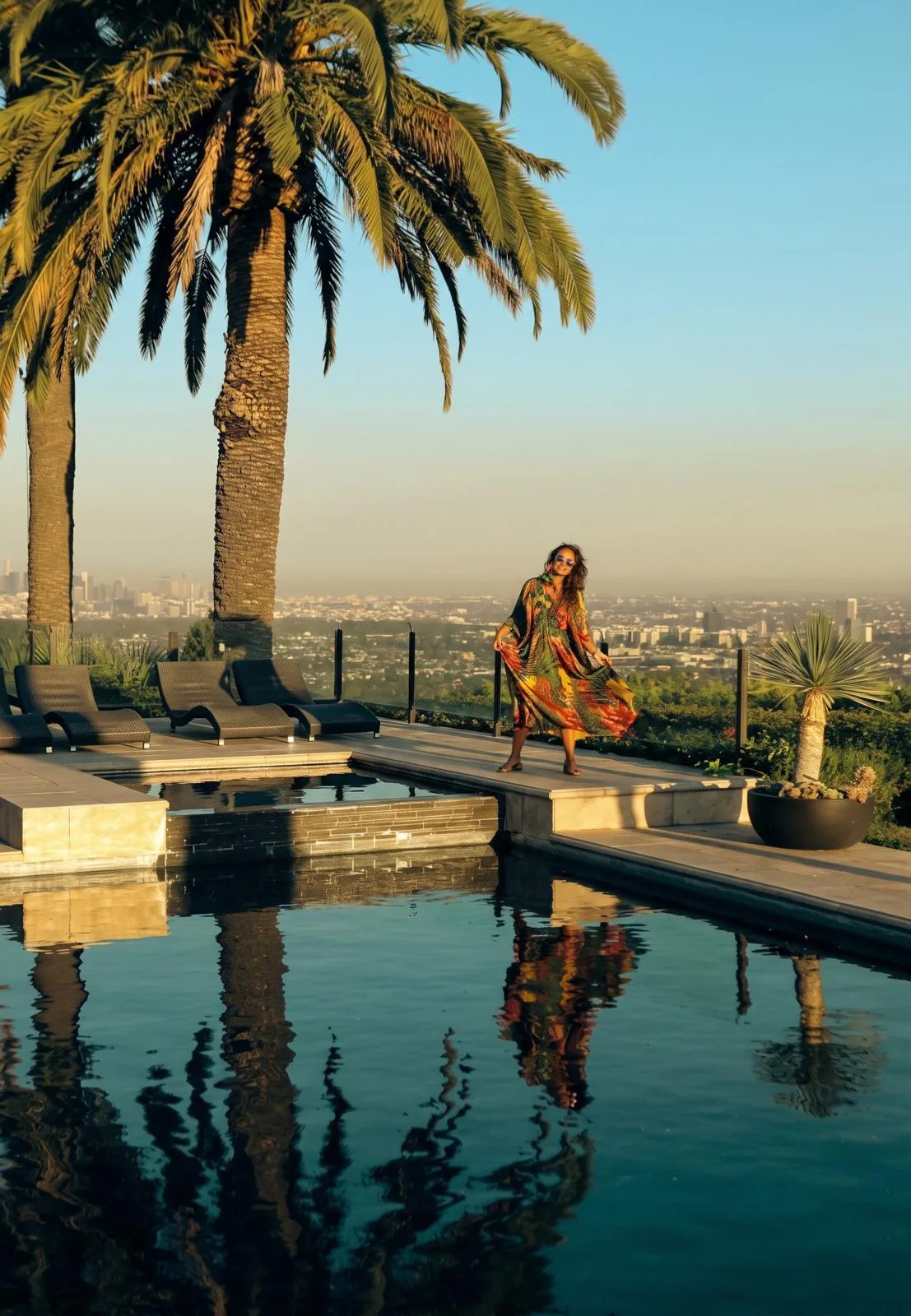 Vanessa Falabella in a flowing dress by a pool lined with palm trees, the city skyline behind her and her reflection in the water