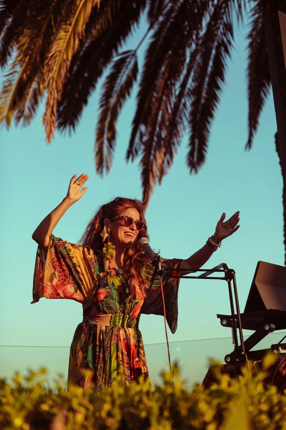 Vanessa Falabella singing at the microphone with both arms raised, under palm trees at golden hour